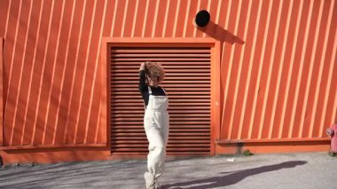 Şık ve kendine güvenen kıvırcık saçlı genç bir kadın canlı ve parlak turuncu bir duvarın önünde duruyor. Modern kıyafetini ve modaya uygun görünümünü göstererek.