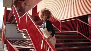 Canlı, kıvırcık saçlı genç bir kadın neşeli kırmızı merdivenlerden neşeyle dans ediyor. Heyecan verici bir şehir ortamında eşsiz stilini ve doğal karizmasını sergiliyor. Olumlu ve eğlenceli.