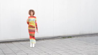 Çarpıcı bir model, hareketleriyle minimalist bir zemin üzerinde zarif bir şekilde dans ederken canlı ve renkli bir elbise sergiliyor, tam anlamıyla neşe, yaratıcılık ve sanatsal ifadeyi somutlaştırıyor.