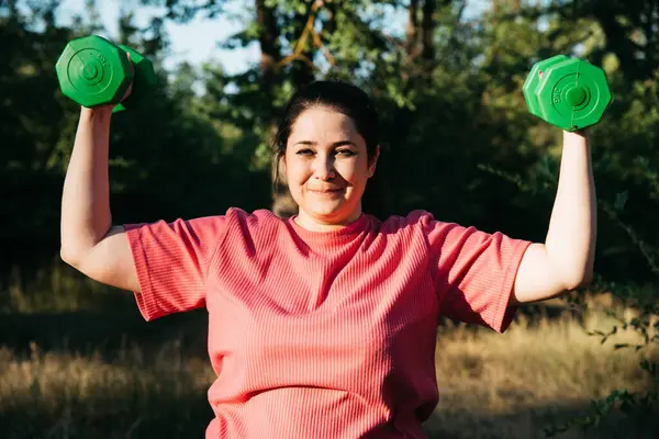 A plus-size woman doing dumbbell exercises outdoors. Outdoor sports. Green dumbbells in the athletes hands. High quality photo