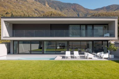 İki katlı modern bir ev, iki iskemle ve beyaz koltuklu küçük bir salon. Önünde güzel yeşil bir çimen var. İçeride kimse yok.