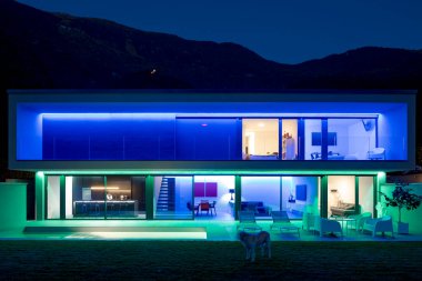Gece havuzu ve bahçesi renkli LED ışıklarıyla aydınlatılmış modern evin ön manzarası. Evin arkasında ormanı olan bir tepe var. İçeride kimse yok.