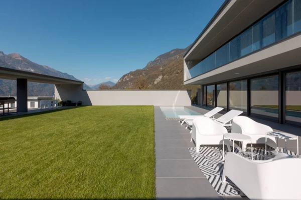 现代化的房子 有修剪过的绿色花园 阴凉的庭院 游泳池 有两张甲板椅和一个白色扶手椅的小休息厅 瑞士阿尔卑斯山的景色 里面没有人 — 图库照片