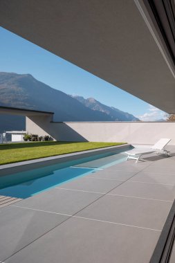 Modern villa bahçesi, yüzme havuzu ve dinlenmek için iskemle. İsviçre Alplerinde güneşli bir gün. İçeride kimse yok.