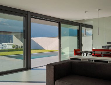 Büyük finistre ile iç modern villa oturma odası. Dışarıda bir yüzme havuzu var. İsviçre Alplerinde güneşli bir gün. İçeride kimse yok.