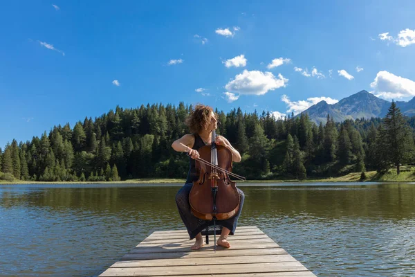 Polish woman and musician plays her cello on her Swiss territory. Sitting in a lei chair on a dock with a mountain lake behind it
