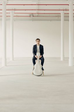 Genç bir çocuk gitar kılıfıyla bir taburede oturuyor. Yenilenmiş eski bir fabrikanın içinde..