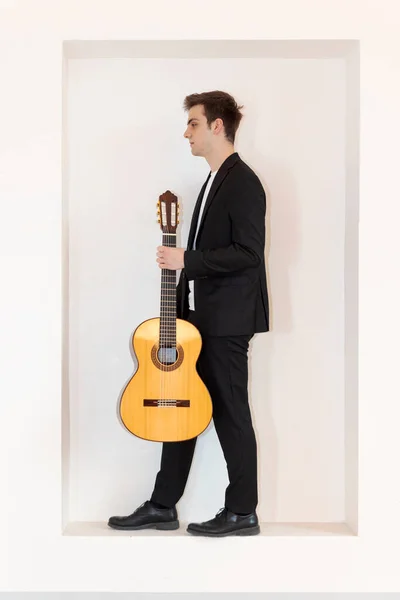 エレガントなスーツの少年が壁のニッチに立っている 彼は片手でギターを持っている 白地に隔離された — ストック写真