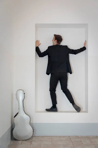 一个穿着雅致西服的年轻人正站在靠墙的架子上 他旁边是他的吉他盒 因白人背景而被隔离 — 图库照片