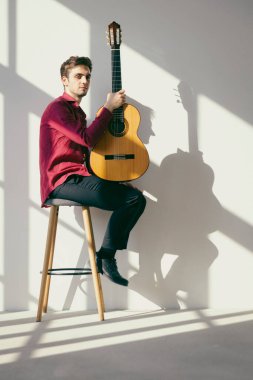 Genç bir çocuk bir eli bacağında gitarını tutarak taburede oturuyor. O, beyaz bir gömlek ve kırmızı bir gömlek giyiyor. Yenilenmiş eski bir fabrikanın içinde..