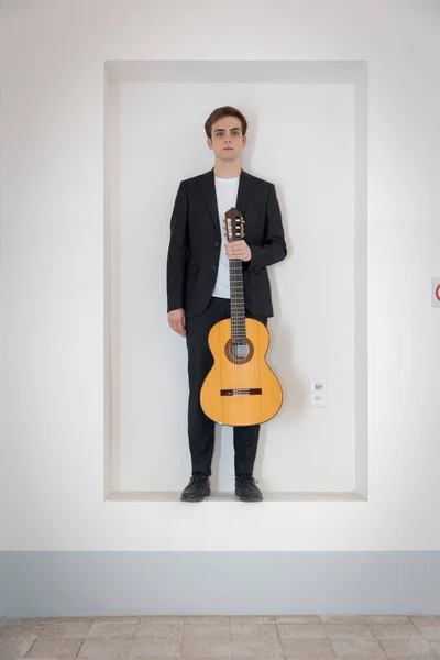 エレガントなスーツの少年が壁のニッチに立っている 一方で彼はギターを持っている 白地に隔離された — ストック写真