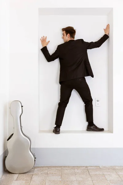 エレガントなスーツを着た若い男が壁に向かってフレームの中に立っている 彼の隣には彼のギターケースがある 白地に隔離された — ストック写真