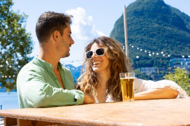 Beyaz dişli güzel, gülümseyen kız arkadaşıyla şakalaşıyor. Arka planda göl ve Lugano 'daki San Salvatore dağı var. Mükemmel bir çift. Masada tek bir bira.