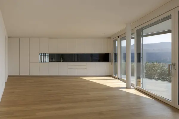 Modern living room with open space and a black and white designed kitchen situated at the end. Large windows allow plenty of sunlight to enter. The room features a parquet floor. Nobody inside
