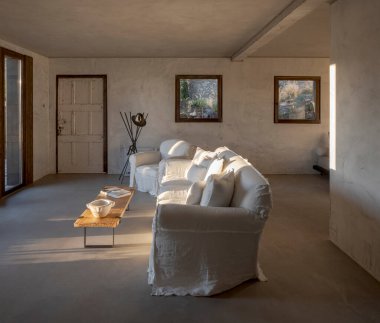 Kanepesi ve koltuğu olan modern bir oturma odası. Sağ taraftan batan güneşin ışığı giriyor. Tasarımcı evi, sert beton duvarlar. İçeride kimse yok.