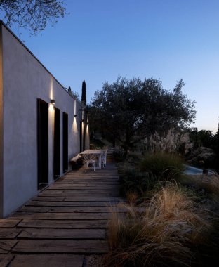 Geceleri aydınlık ışıkları, ahşap bir yürüyüş yolu ve koltukları olan bir masası olan modern bir kırsal konutun dışında. Etrafı yeşillik ve zeytin ağaçlarıyla çevrili. İçeride kimse yok.
