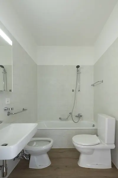 Interior of a bathroom in a normal flat, neither poor nor rich. Everything is white and there are no people inside. The floor is made of wood