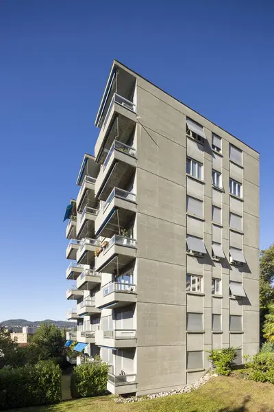 Exterior view of an old rough concrete apartment building with balconies and blinds. There is a green garden and trees around. No one inside.