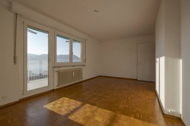 Boş parke döşemeli oda, pencerenin altında büyük bir radyatör var. Kapıdan, küçük balkona çıkabilirsiniz. İçeride kimse yok.