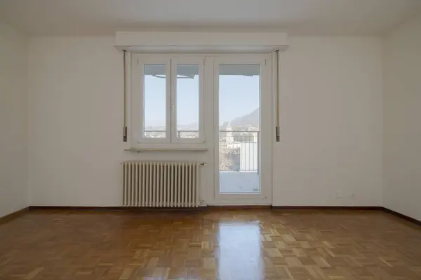 Empty living room with a bright window looking out at the blue sky. An old radiator hangs on the white walls. No one inside