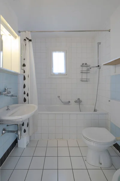Old bathroom with white tiles, at the end there is a bathtub with shower and open curtain. There is also a small window. Nobody inside