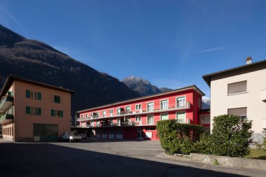 Aşağıda park alanı olan kırmızı parmaklıklı ev. Bazı pencereler açık, diğerleri yeşil panjurlarla kapatılmış. Mavi gökyüzü ve arkadaki dağlar. İçeride kimse yok.