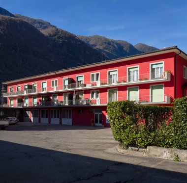 Aşağıda park alanı olan kırmızı parmaklıklı ev. Bazı pencereler açık, diğerleri yeşil panjurlarla kapatılmış. Mavi gökyüzü ve arkadaki dağlar. İçeride kimse yok.