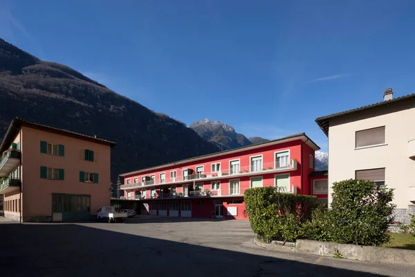 Aşağıda park alanı olan kırmızı parmaklıklı ev. Bazı pencereler açık, diğerleri yeşil panjurlarla kapatılmış. Mavi gökyüzü ve arkadaki dağlar. İçeride kimse yok.