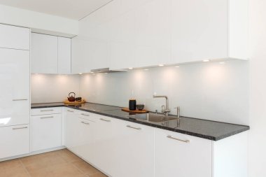 Parlak beyaz, minimalist, siyah mermer tezgahlı modern mutfak. Sahne ışıkları tezgahı aydınlatıyor, üzerinde iki tepsi var, biri çaydanlık, diğeri vazo. İçeride kimse yok.