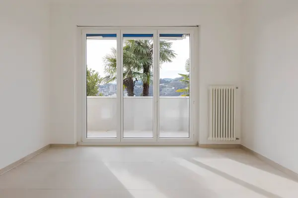 空荡荡的房间 后面有一个大窗户 通往阳台 在白墙的右边可以看到散热器或加热器 图库图片