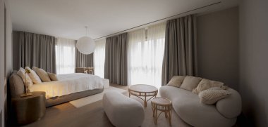 Modern bir evde minimalist tasarım yatak odası panoraması. Komodinli büyük bir çift kişilik yatak, hafif bir kanepe ve sandalyeli bir masa var. İçeride kimse yok.