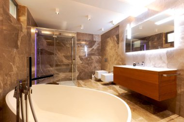 Mermer duvarları ve zemini olan modern bir banyo. Odanın ortasında bir küvet. Cam duvarlı duş ve üzerinde ayna olan lavabo. İçeride kimse yok.