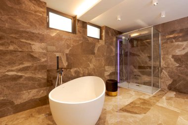 Mermer duvarları ve zemini olan modern bir banyo. Odanın ortasında bir küvet. Arka planda cam duvarlı bir duş görebilirsiniz. İçeride kimse yok.