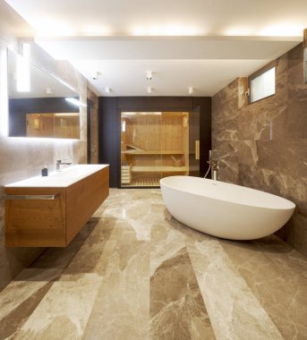 Mermer duvarları ve zemini olan modern bir banyo. Odanın ortasında bir küvet. Arka planda cam kapılı bir sauna görebilirsiniz. İçeride kimse yok.