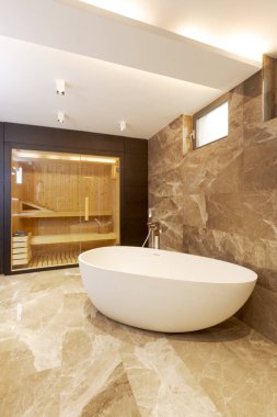 Mermer duvarları ve zemini olan modern bir banyo. Odanın ortasında bir küvet. Arka planda cam kapılı bir sauna görebilirsiniz. İçeride kimse yok.