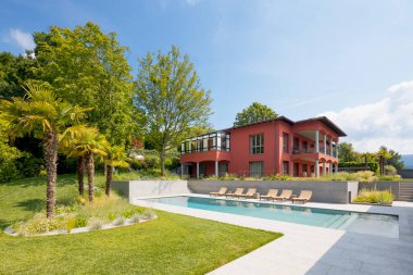 Ağaçları olan büyük yeşil bir bahçedeki kırmızı duvarlı villa. Büyük bir yüzme havuzu ve dinlenme yerleri var. Güneşli yaz günü.