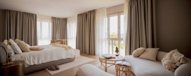 Modern bir evde minimalist tasarım yatak odası panoraması. Komodinli büyük bir çift kişilik yatak, hafif bir kanepe ve sandalyeli bir masa var. İçeride kimse yok.