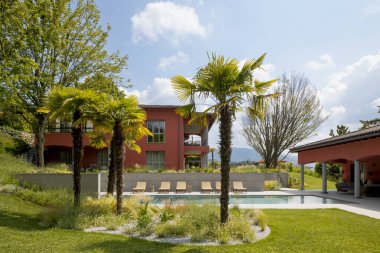 Modern bir villa manzarası, yüzme havuzu, şezlong ve büyük bir bahçesi var. Ön planda palmiye ağaçları var, içinde kimse yok.