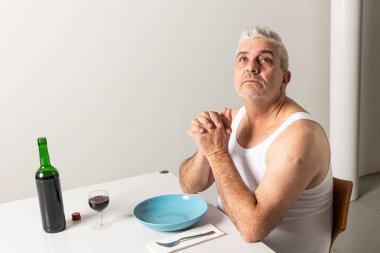 Portrait of an Italian man in a white undershirt sitting at a table while waiting for the pasta to be ready and praying while looking upwards. Isolated scene of Italian domestic life clipart