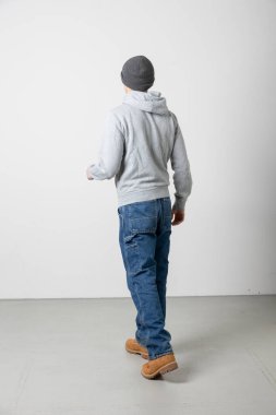 Arkadan beyaz bir arka planda tek parça halinde duran genç bir çocuk. Tüm vücudunu görebilirsin. Kot pantolon ve kazak giyiyor.. 