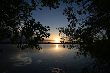 Gün batımı ağaçların arasından gölün üzerinde