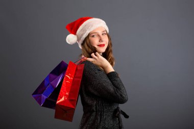 Noel hediyesi almak için alışveriş yapan bir kadın. Kafkasyalı genç bir kız alışveriş torbaları ve Noel Baba şapkasıyla gülümsüyor. Uzayı kenarda kopyala, gri arkaplanda izole et..