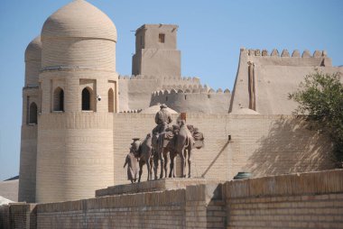 Özbekistan 'ın başkenti Khiva' nın tuğla ve toprak duvarları ve kubbeli gözetleme kulelerinin önünde, İpek Yolu 'nda gezginlerin, develerin ve dromedarların bulunduğu bir çöl karavan bronz heykeli.