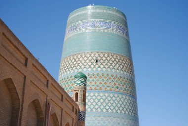 Özbekistan 'ın başkenti Khiva' da, kubbe kulesi, tuğla süslemeli duvarları ve mavi gökyüzü olan bir binanın arkasında zengin boyanmış fayanslara ve mozaiğe sahip Kalta Minor minaresi.