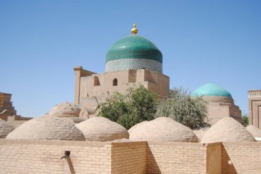 Zirvesinde altın küreler olan fayanslı bir kubbe ve zengin boyanmış kiremitlerle süslü duvarlar ve Özbekistan 'ın başkenti Khiva' da zeytin ağaçları ve mavi gökyüzü bulunan toprak tuğlaları.