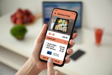 female hands holding phone with app delivery food on the screen over a table with laptop in room
