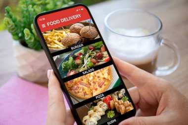Kadın elleri yemek dağıtım uygulaması olan telefonu ekrandaki masanın arka planında tutuyor.