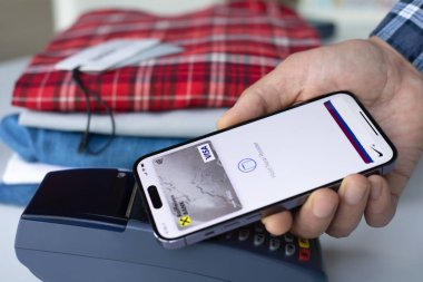 Alanya, Türkiye - 16 Nisan 2024: Apple Pay Raiffeisenbank uygulamalı iPhone 15 Pro Max ile el ele tutuşmak ve mağazadan POS terminal kıyafetleri satın almak.
