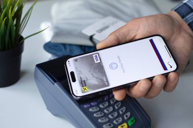 Alanya, Türkiye - 16 Nisan 2024: Apple iPhone 15 Pro Max 'i Apple Pay Raiffeisenbank uygulaması ile elinde tutan ve mağazadan POS terminal kıyafetleri satın alan adam.