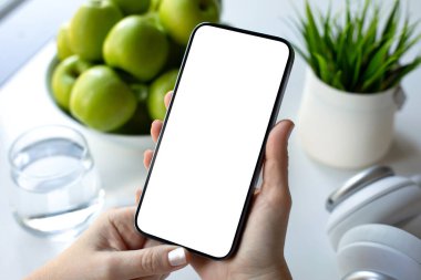 Kadın elleri, kafede yeşil elma ve kulaklıklı bir masanın arka planında izole bir ekranla telefonu tutuyor.